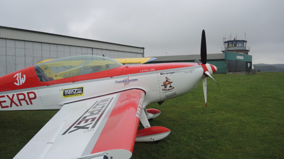 Ein rot-weiß lackiertes Kunstflugzeug steht auf einer Rasenfläche vor einem Hangar und dem Tower eines kleinen Flugplatzes. Der Propeller ruht, während der Himmel von tief hängenden grauen Wolken bedeckt ist. Die Aufnahme zeigt das Flugzeug aus seitlicher Perspektive und vermittelt die ruhige Atmosphäre eines Flugplatzes vor dem Start oder nach der Landung.