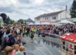 Bei einem Feuerwehrfest führen Kinder der Kinderfeuerwehr vor großem Publikum eine Löschübung vor. Mit Schutzkleidung und Helmen richten sie Wasserstrahlen auf ein Übungshaus, während zahlreiche Familien und Besucher die Vorführung entlang der abgesperrten Straße verfolgen. Im Hintergrund sind Feuerwehrfahrzeuge, Zelte und Feststände zu sehen, über der Szene ziehen dunkle Wolken auf.