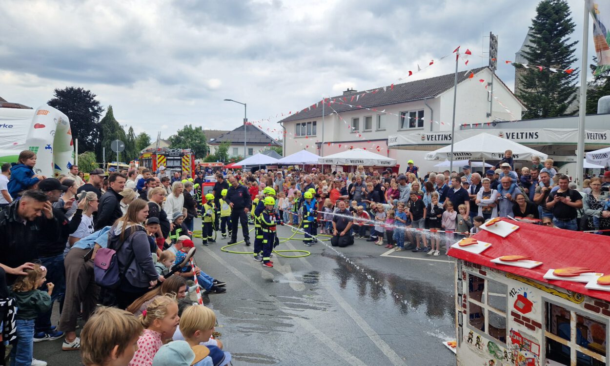 Bei einem Feuerwehrfest führen Kinder der Kinderfeuerwehr vor großem Publikum eine Löschübung vor. Mit Schutzkleidung und Helmen richten sie Wasserstrahlen auf ein Übungshaus, während zahlreiche Familien und Besucher die Vorführung entlang der abgesperrten Straße verfolgen. Im Hintergrund sind Feuerwehrfahrzeuge, Zelte und Feststände zu sehen, über der Szene ziehen dunkle Wolken auf.