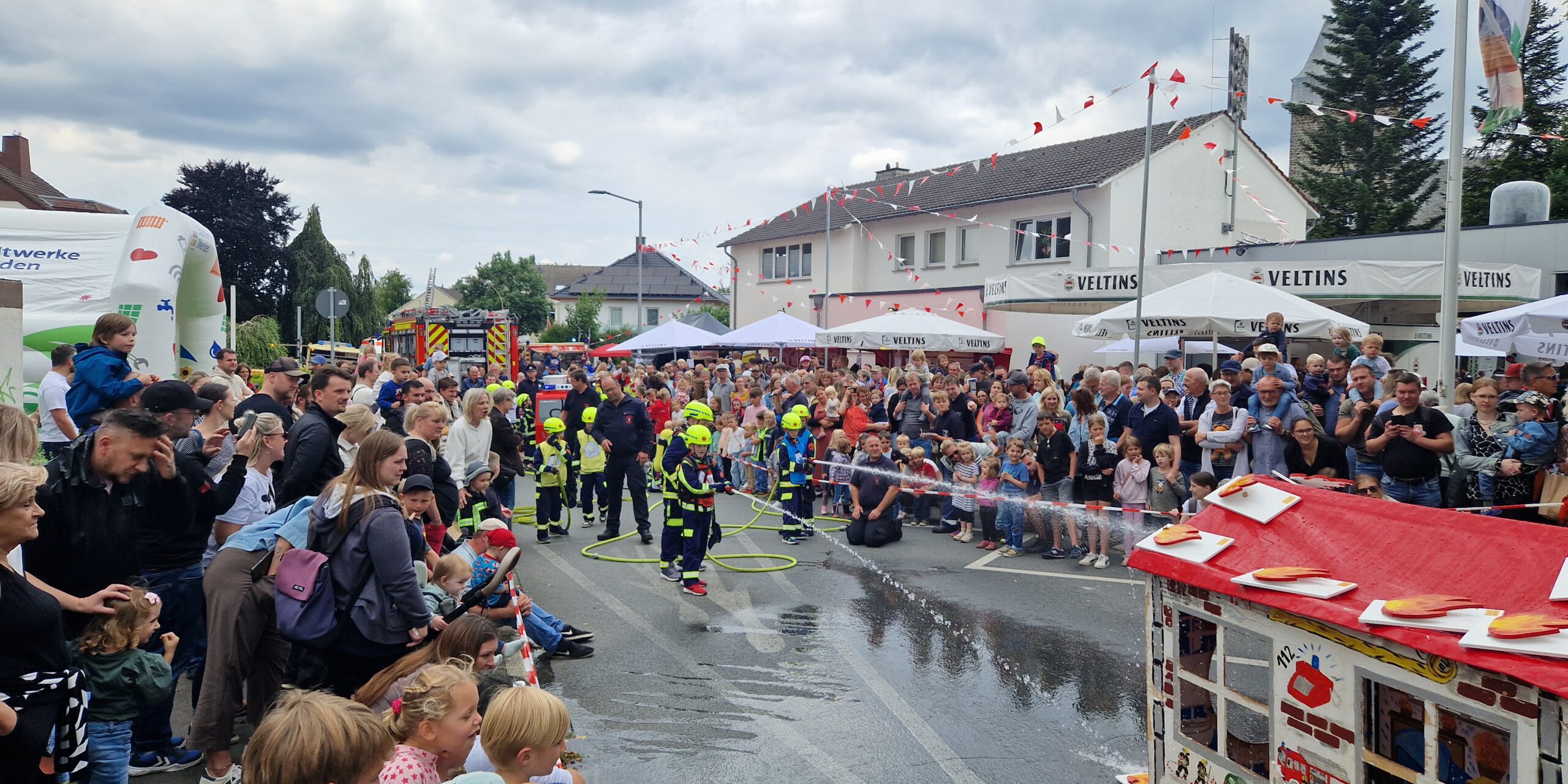 Bei einem Feuerwehrfest führen Kinder der Kinderfeuerwehr vor großem Publikum eine Löschübung vor. Mit Schutzkleidung und Helmen richten sie Wasserstrahlen auf ein Übungshaus, während zahlreiche Familien und Besucher die Vorführung entlang der abgesperrten Straße verfolgen. Im Hintergrund sind Feuerwehrfahrzeuge, Zelte und Feststände zu sehen, über der Szene ziehen dunkle Wolken auf.