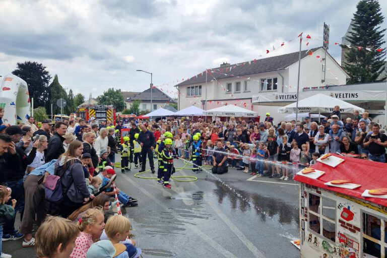 Bei einem Feuerwehrfest führen Kinder der Kinderfeuerwehr vor großem Publikum eine Löschübung vor. Mit Schutzkleidung und Helmen richten sie Wasserstrahlen auf ein Übungshaus, während zahlreiche Familien und Besucher die Vorführung entlang der abgesperrten Straße verfolgen. Im Hintergrund sind Feuerwehrfahrzeuge, Zelte und Feststände zu sehen, über der Szene ziehen dunkle Wolken auf.