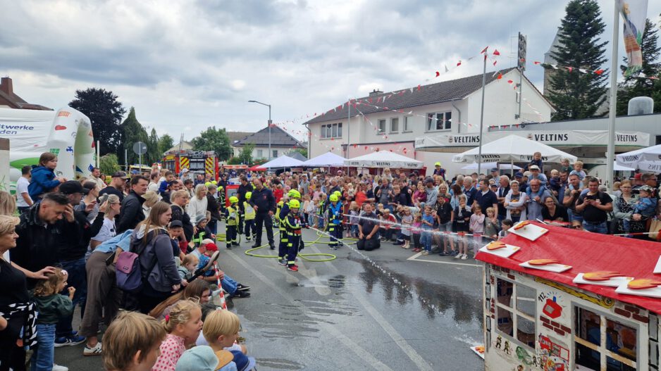 Bei einem Feuerwehrfest führen Kinder der Kinderfeuerwehr vor großem Publikum eine Löschübung vor. Mit Schutzkleidung und Helmen richten sie Wasserstrahlen auf ein Übungshaus, während zahlreiche Familien und Besucher die Vorführung entlang der abgesperrten Straße verfolgen. Im Hintergrund sind Feuerwehrfahrzeuge, Zelte und Feststände zu sehen, über der Szene ziehen dunkle Wolken auf.
