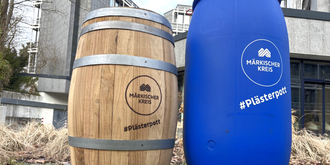 Zwei Regentonnen stehen auf einer Grünfläche vor einem Verwaltungsgebäude. Links ist eine Tonne in Holzfass-Optik, rechts eine blaue Kunststoff-Regentonne zu sehen. Beide tragen das Logo des Märkischen Kreises sowie den Schriftzug „#Plästerpott“. Die Aufnahme veranschaulicht zwei unterschiedliche Modelle von Regentonnen zur Regenwassernutzung.