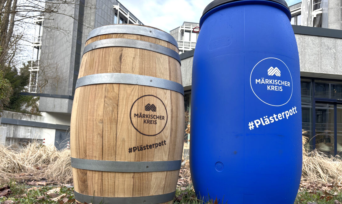 Zwei Regentonnen stehen auf einer Grünfläche vor einem Verwaltungsgebäude. Links ist eine Tonne in Holzfass-Optik, rechts eine blaue Kunststoff-Regentonne zu sehen. Beide tragen das Logo des Märkischen Kreises sowie den Schriftzug „#Plästerpott“. Die Aufnahme veranschaulicht zwei unterschiedliche Modelle von Regentonnen zur Regenwassernutzung.