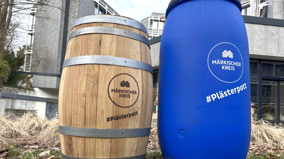 Zwei Regentonnen stehen auf einer Grünfläche vor einem Verwaltungsgebäude. Links ist eine Tonne in Holzfass-Optik, rechts eine blaue Kunststoff-Regentonne zu sehen. Beide tragen das Logo des Märkischen Kreises sowie den Schriftzug „#Plästerpott“. Die Aufnahme veranschaulicht zwei unterschiedliche Modelle von Regentonnen zur Regenwassernutzung.