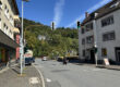 Straßenkreuzung in Altena bei sonnigem Wetter mit mehreren Ampelanlagen. Autos und ein Motorrad fahren über die Straße, im Hintergrund ist ein Linienbus an einer Haltestelle zu sehen. Wohnhäuser säumen die Straße, dahinter steigt ein dicht bewaldeter Hang unter blauem Himmel an.