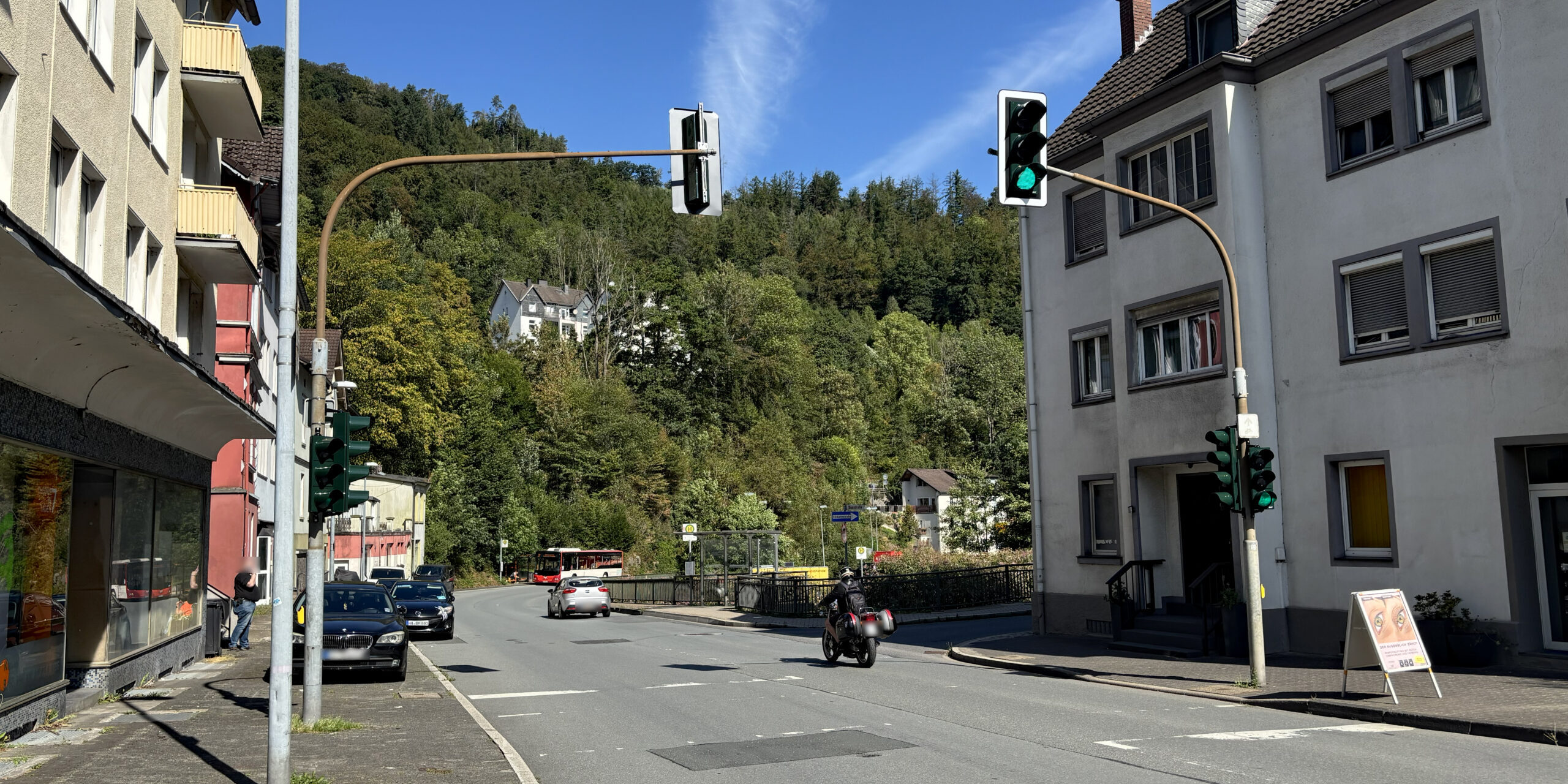 Straßenkreuzung in Altena bei sonnigem Wetter mit mehreren Ampelanlagen. Autos und ein Motorrad fahren über die Straße, im Hintergrund ist ein Linienbus an einer Haltestelle zu sehen. Wohnhäuser säumen die Straße, dahinter steigt ein dicht bewaldeter Hang unter blauem Himmel an.