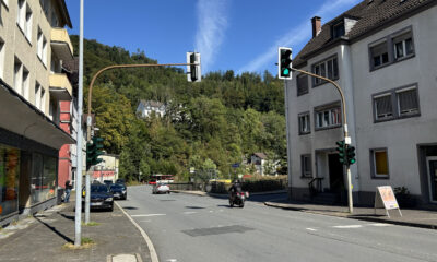 Straßenkreuzung in Altena bei sonnigem Wetter mit mehreren Ampelanlagen. Autos und ein Motorrad fahren über die Straße, im Hintergrund ist ein Linienbus an einer Haltestelle zu sehen. Wohnhäuser säumen die Straße, dahinter steigt ein dicht bewaldeter Hang unter blauem Himmel an.