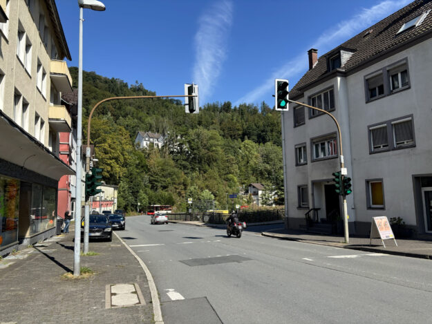 Foto: Stadt Altena Straßenkreuzung in Altena bei sonnigem Wetter mit mehreren Ampelanlagen. Autos und ein Motorrad fahren über die Straße, im Hintergrund ist ein Linienbus an einer Haltestelle zu sehen. Wohnhäuser säumen die Straße, dahinter steigt ein dicht bewaldeter Hang unter blauem Himmel an.
