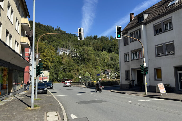 Foto: Stadt Altena Straßenkreuzung in Altena bei sonnigem Wetter mit mehreren Ampelanlagen. Autos und ein Motorrad fahren über die Straße, im Hintergrund ist ein Linienbus an einer Haltestelle zu sehen. Wohnhäuser säumen die Straße, dahinter steigt ein dicht bewaldeter Hang unter blauem Himmel an.
