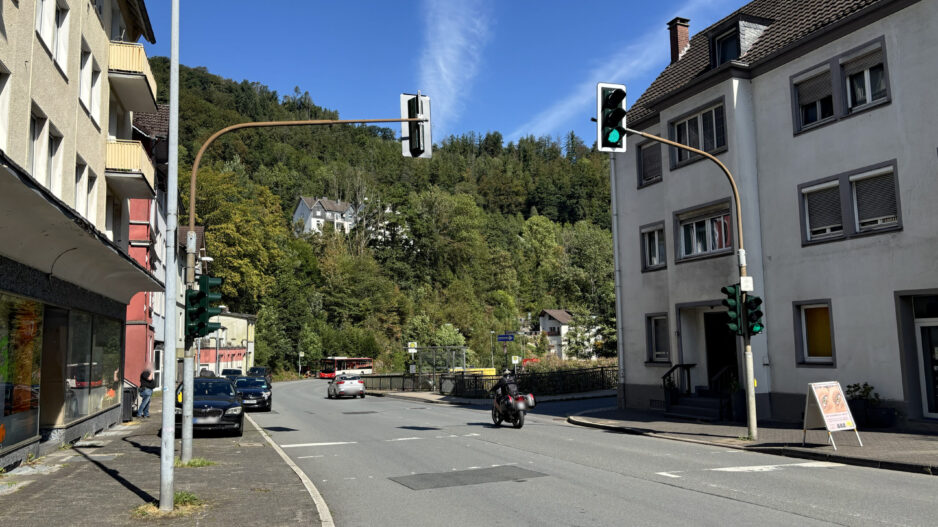 Straßenkreuzung in Altena bei sonnigem Wetter mit mehreren Ampelanlagen. Autos und ein Motorrad fahren über die Straße, im Hintergrund ist ein Linienbus an einer Haltestelle zu sehen. Wohnhäuser säumen die Straße, dahinter steigt ein dicht bewaldeter Hang unter blauem Himmel an.