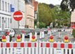Rot-weiße Absperrgitter und Leitbaken sperren eine Fahrspur einer innerstädtischen Straße wegen einer Baustelle. Der Verkehr wird einspurig an der Baustelle vorbeigeführt, wodurch sich eine Fahrzeugschlange bildet. Im Hintergrund sind Wohn- und Geschäftshäuser, Ampeln sowie weitere Absperrungen zu sehen, die die Verkehrsführung durch den Baustellenbereich kennzeichnen.