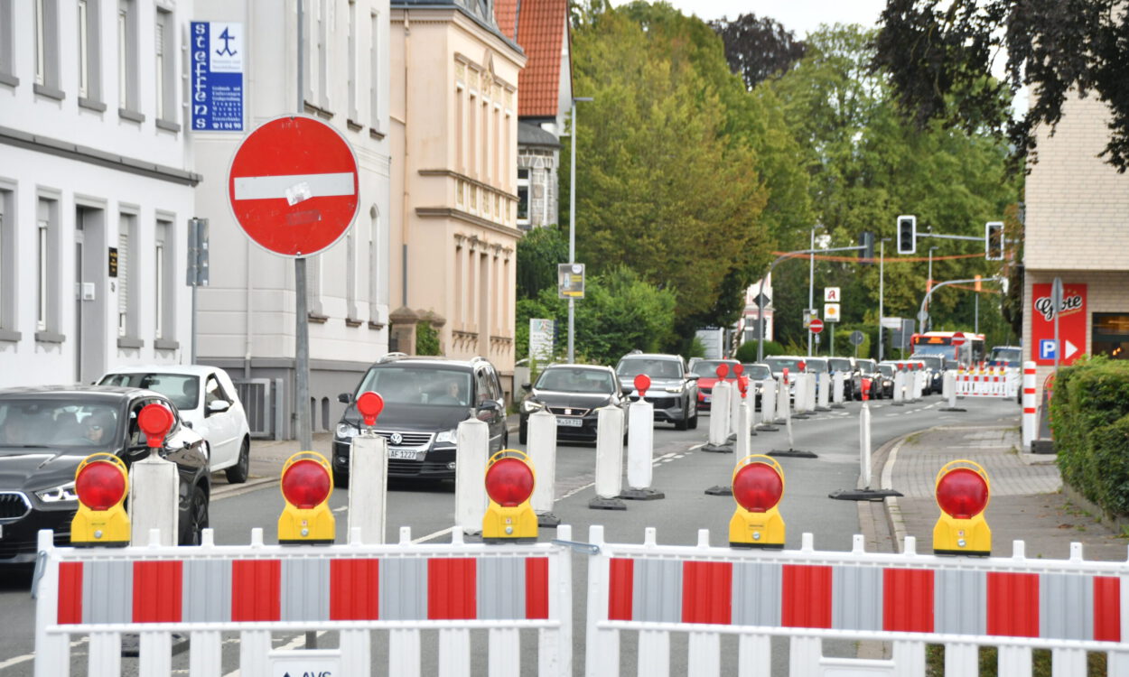 Rot-weiße Absperrgitter und Leitbaken sperren eine Fahrspur einer innerstädtischen Straße wegen einer Baustelle. Der Verkehr wird einspurig an der Baustelle vorbeigeführt, wodurch sich eine Fahrzeugschlange bildet. Im Hintergrund sind Wohn- und Geschäftshäuser, Ampeln sowie weitere Absperrungen zu sehen, die die Verkehrsführung durch den Baustellenbereich kennzeichnen.