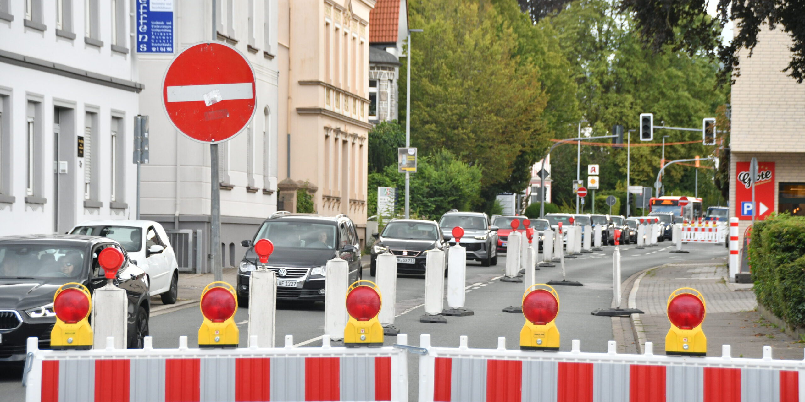 Rot-weiße Absperrgitter und Leitbaken sperren eine Fahrspur einer innerstädtischen Straße wegen einer Baustelle. Der Verkehr wird einspurig an der Baustelle vorbeigeführt, wodurch sich eine Fahrzeugschlange bildet. Im Hintergrund sind Wohn- und Geschäftshäuser, Ampeln sowie weitere Absperrungen zu sehen, die die Verkehrsführung durch den Baustellenbereich kennzeichnen.