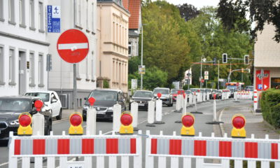 Rot-weiße Absperrgitter und Leitbaken sperren eine Fahrspur einer innerstädtischen Straße wegen einer Baustelle. Der Verkehr wird einspurig an der Baustelle vorbeigeführt, wodurch sich eine Fahrzeugschlange bildet. Im Hintergrund sind Wohn- und Geschäftshäuser, Ampeln sowie weitere Absperrungen zu sehen, die die Verkehrsführung durch den Baustellenbereich kennzeichnen.