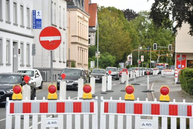Rot-weiße Absperrgitter und Leitbaken sperren eine Fahrspur einer innerstädtischen Straße wegen einer Baustelle. Der Verkehr wird einspurig an der Baustelle vorbeigeführt, wodurch sich eine Fahrzeugschlange bildet. Im Hintergrund sind Wohn- und Geschäftshäuser, Ampeln sowie weitere Absperrungen zu sehen, die die Verkehrsführung durch den Baustellenbereich kennzeichnen.