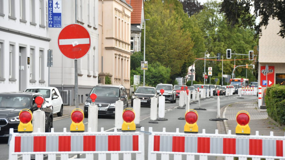 Rot-weiße Absperrgitter und Leitbaken sperren eine Fahrspur einer innerstädtischen Straße wegen einer Baustelle. Der Verkehr wird einspurig an der Baustelle vorbeigeführt, wodurch sich eine Fahrzeugschlange bildet. Im Hintergrund sind Wohn- und Geschäftshäuser, Ampeln sowie weitere Absperrungen zu sehen, die die Verkehrsführung durch den Baustellenbereich kennzeichnen.