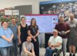 Acht Personen stehen und sitzen gemeinsam in einem Besprechungsraum vor einem großen Bildschirm mit einer digitalen Stadtkarte. Im Mittelpunkt steht die Vorstellung eines Dashboards zur Erfassung von Behindertenparkplätzen. Im Hintergrund sind Informationsposter und eine großformatige Luftaufnahme der Stadt mit grafischen Vernetzungssymbolen zu sehen. Ein Rollstuhlfahrer ist Teil der Gruppe und unterstreicht den Bezug zum Thema Barrierefreiheit.