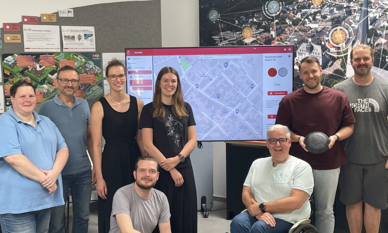 Acht Personen stehen und sitzen gemeinsam in einem Besprechungsraum vor einem großen Bildschirm mit einer digitalen Stadtkarte. Im Mittelpunkt steht die Vorstellung eines Dashboards zur Erfassung von Behindertenparkplätzen. Im Hintergrund sind Informationsposter und eine großformatige Luftaufnahme der Stadt mit grafischen Vernetzungssymbolen zu sehen. Ein Rollstuhlfahrer ist Teil der Gruppe und unterstreicht den Bezug zum Thema Barrierefreiheit.