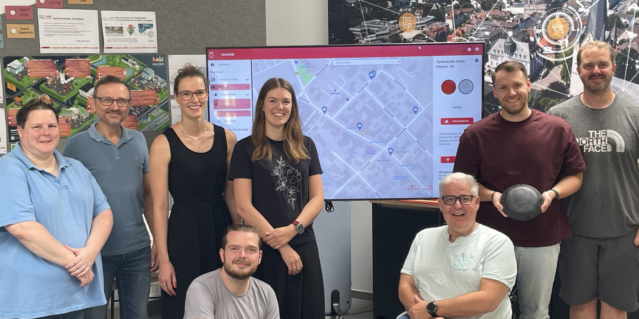 Acht Personen stehen und sitzen gemeinsam in einem Besprechungsraum vor einem großen Bildschirm mit einer digitalen Stadtkarte. Im Mittelpunkt steht die Vorstellung eines Dashboards zur Erfassung von Behindertenparkplätzen. Im Hintergrund sind Informationsposter und eine großformatige Luftaufnahme der Stadt mit grafischen Vernetzungssymbolen zu sehen. Ein Rollstuhlfahrer ist Teil der Gruppe und unterstreicht den Bezug zum Thema Barrierefreiheit.