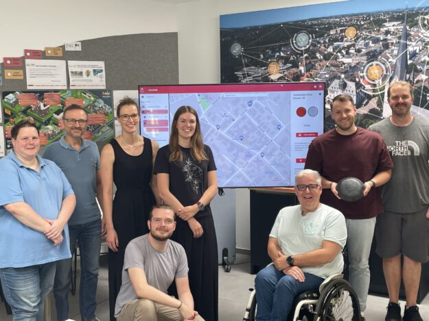 Foto: M. Collard/Stadt Menden Acht Personen stehen und sitzen gemeinsam in einem Besprechungsraum vor einem großen Bildschirm mit einer digitalen Stadtkarte. Im Mittelpunkt steht die Vorstellung eines Dashboards zur Erfassung von Behindertenparkplätzen. Im Hintergrund sind Informationsposter und eine großformatige Luftaufnahme der Stadt mit grafischen Vernetzungssymbolen zu sehen. Ein Rollstuhlfahrer ist Teil der Gruppe und unterstreicht den Bezug zum Thema Barrierefreiheit.