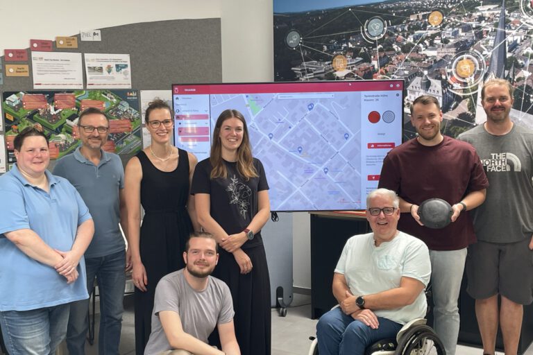 Acht Personen stehen und sitzen gemeinsam in einem Besprechungsraum vor einem großen Bildschirm mit einer digitalen Stadtkarte. Im Mittelpunkt steht die Vorstellung eines Dashboards zur Erfassung von Behindertenparkplätzen. Im Hintergrund sind Informationsposter und eine großformatige Luftaufnahme der Stadt mit grafischen Vernetzungssymbolen zu sehen. Ein Rollstuhlfahrer ist Teil der Gruppe und unterstreicht den Bezug zum Thema Barrierefreiheit.