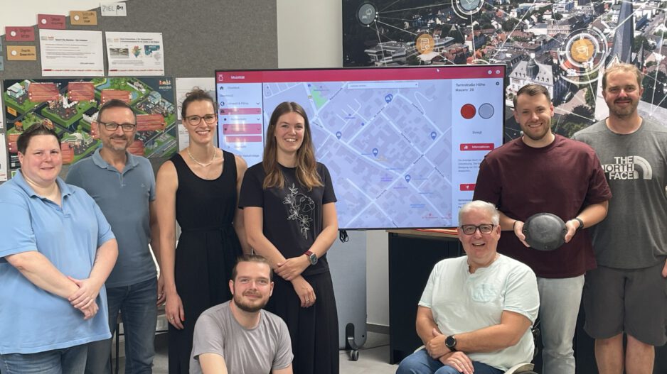 Acht Personen stehen und sitzen gemeinsam in einem Besprechungsraum vor einem großen Bildschirm mit einer digitalen Stadtkarte. Im Mittelpunkt steht die Vorstellung eines Dashboards zur Erfassung von Behindertenparkplätzen. Im Hintergrund sind Informationsposter und eine großformatige Luftaufnahme der Stadt mit grafischen Vernetzungssymbolen zu sehen. Ein Rollstuhlfahrer ist Teil der Gruppe und unterstreicht den Bezug zum Thema Barrierefreiheit.