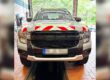Ein silberner Pick-up steht in einer Fahrzeughalle auf einer Hebebühne oder über einer Wartungsgrube. Auf der Motorhaube sind rot-weiße Warnmarkierungen angebracht, auf dem Dach befindet sich eine LED-Lichtleiste. Das Fahrzeug wirkt für den Einsatz im Straßenunterhalt oder im kommunalen Dienst ausgerüstet. Durch das geöffnete Hallentor ist im Hintergrund ein bewaldeter Bereich zu sehen.