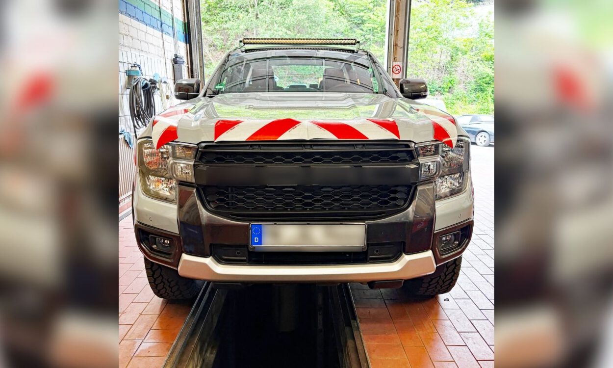 Ein silberner Pick-up steht in einer Fahrzeughalle auf einer Hebebühne oder über einer Wartungsgrube. Auf der Motorhaube sind rot-weiße Warnmarkierungen angebracht, auf dem Dach befindet sich eine LED-Lichtleiste. Das Fahrzeug wirkt für den Einsatz im Straßenunterhalt oder im kommunalen Dienst ausgerüstet. Durch das geöffnete Hallentor ist im Hintergrund ein bewaldeter Bereich zu sehen.