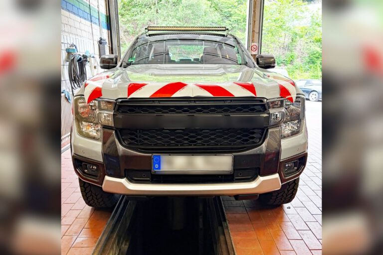 Ein silberner Pick-up steht in einer Fahrzeughalle auf einer Hebebühne oder über einer Wartungsgrube. Auf der Motorhaube sind rot-weiße Warnmarkierungen angebracht, auf dem Dach befindet sich eine LED-Lichtleiste. Das Fahrzeug wirkt für den Einsatz im Straßenunterhalt oder im kommunalen Dienst ausgerüstet. Durch das geöffnete Hallentor ist im Hintergrund ein bewaldeter Bereich zu sehen.