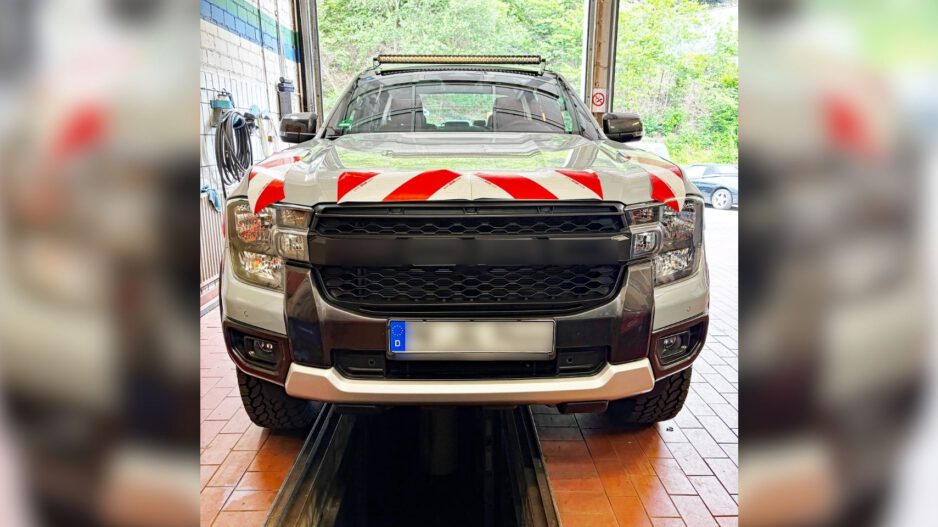 Ein silberner Pick-up steht in einer Fahrzeughalle auf einer Hebebühne oder über einer Wartungsgrube. Auf der Motorhaube sind rot-weiße Warnmarkierungen angebracht, auf dem Dach befindet sich eine LED-Lichtleiste. Das Fahrzeug wirkt für den Einsatz im Straßenunterhalt oder im kommunalen Dienst ausgerüstet. Durch das geöffnete Hallentor ist im Hintergrund ein bewaldeter Bereich zu sehen.