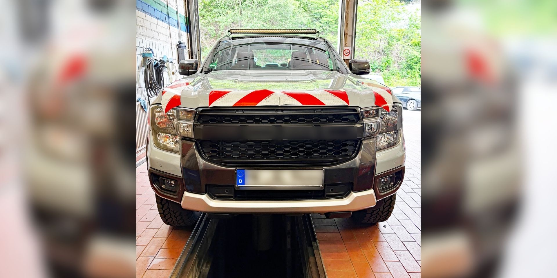 Ein silberner Pick-up steht in einer Fahrzeughalle auf einer Hebebühne oder über einer Wartungsgrube. Auf der Motorhaube sind rot-weiße Warnmarkierungen angebracht, auf dem Dach befindet sich eine LED-Lichtleiste. Das Fahrzeug wirkt für den Einsatz im Straßenunterhalt oder im kommunalen Dienst ausgerüstet. Durch das geöffnete Hallentor ist im Hintergrund ein bewaldeter Bereich zu sehen.