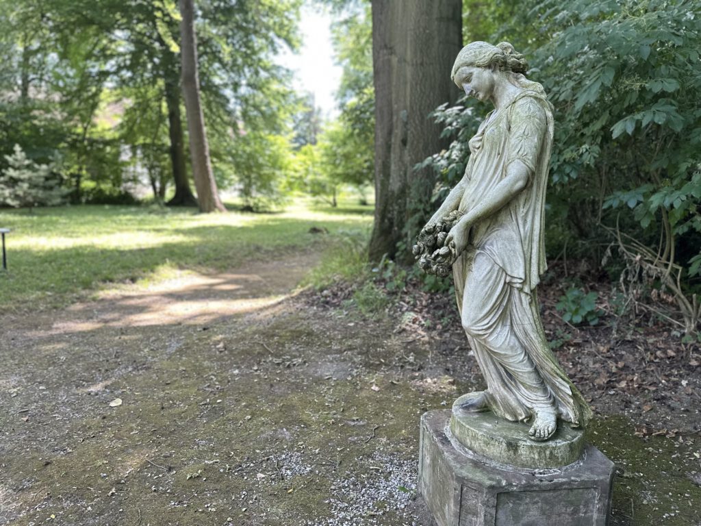 Eine steinerne Frauenfigur mit langem Gewand und einem Blumenkranz in den Händen steht auf einem Sockel am Rand eines geschwungenen Parkwegs. Umgeben von hohen Bäumen und dichtem Grün fügt sich die verwitterte Skulptur harmonisch in die ruhige, parkähnliche Landschaft ein. Das weiche Sonnenlicht und der leicht unscharfe Hintergrund verleihen der Szene eine idyllische, fast märchenhafte Atmosphäre.