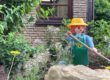 Eine große Spielfigur im Stil einer Gärtnerin mit gelbem Hut, grüner Schürze und langem Gartenwerkzeug steht zwischen Blumen, Sträuchern und Natursteinen in einem liebevoll angelegten Vorgarten. Im Hintergrund sind die Klinkerfassade und ein Fenster eines Wohnhauses zu sehen. Die Szene vermittelt den Eindruck eines gepflegten, naturnah gestalteten Gartens mit dekorativer Figur.