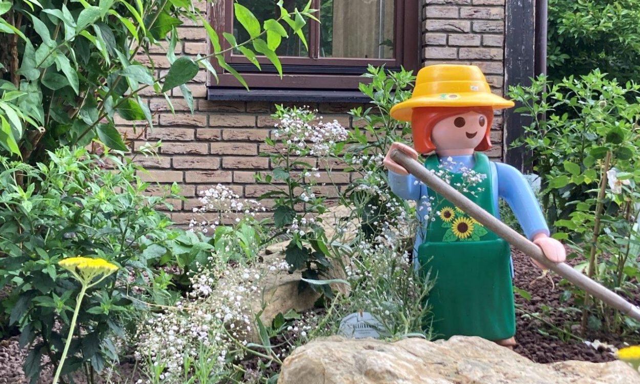 Eine große Spielfigur im Stil einer Gärtnerin mit gelbem Hut, grüner Schürze und langem Gartenwerkzeug steht zwischen Blumen, Sträuchern und Natursteinen in einem liebevoll angelegten Vorgarten. Im Hintergrund sind die Klinkerfassade und ein Fenster eines Wohnhauses zu sehen. Die Szene vermittelt den Eindruck eines gepflegten, naturnah gestalteten Gartens mit dekorativer Figur.