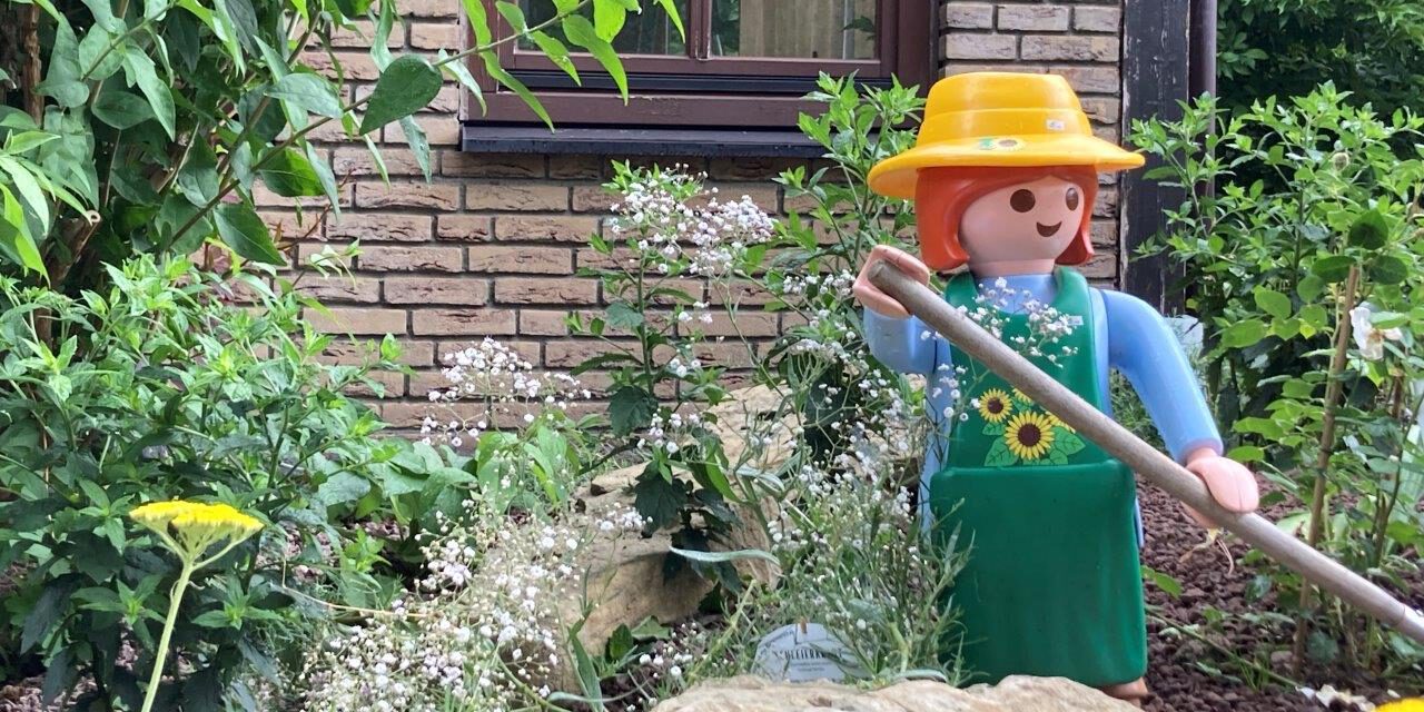 Eine große Spielfigur im Stil einer Gärtnerin mit gelbem Hut, grüner Schürze und langem Gartenwerkzeug steht zwischen Blumen, Sträuchern und Natursteinen in einem liebevoll angelegten Vorgarten. Im Hintergrund sind die Klinkerfassade und ein Fenster eines Wohnhauses zu sehen. Die Szene vermittelt den Eindruck eines gepflegten, naturnah gestalteten Gartens mit dekorativer Figur.