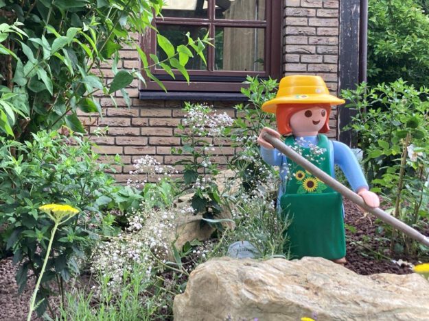 Foto: Thomas Schepp/Stadt Menden Eine große Spielfigur im Stil einer Gärtnerin mit gelbem Hut, grüner Schürze und langem Gartenwerkzeug steht zwischen Blumen, Sträuchern und Natursteinen in einem liebevoll angelegten Vorgarten. Im Hintergrund sind die Klinkerfassade und ein Fenster eines Wohnhauses zu sehen. Die Szene vermittelt den Eindruck eines gepflegten, naturnah gestalteten Gartens mit dekorativer Figur.