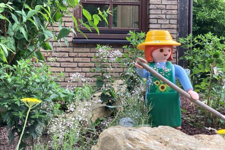 Eine große Spielfigur im Stil einer Gärtnerin mit gelbem Hut, grüner Schürze und langem Gartenwerkzeug steht zwischen Blumen, Sträuchern und Natursteinen in einem liebevoll angelegten Vorgarten. Im Hintergrund sind die Klinkerfassade und ein Fenster eines Wohnhauses zu sehen. Die Szene vermittelt den Eindruck eines gepflegten, naturnah gestalteten Gartens mit dekorativer Figur.