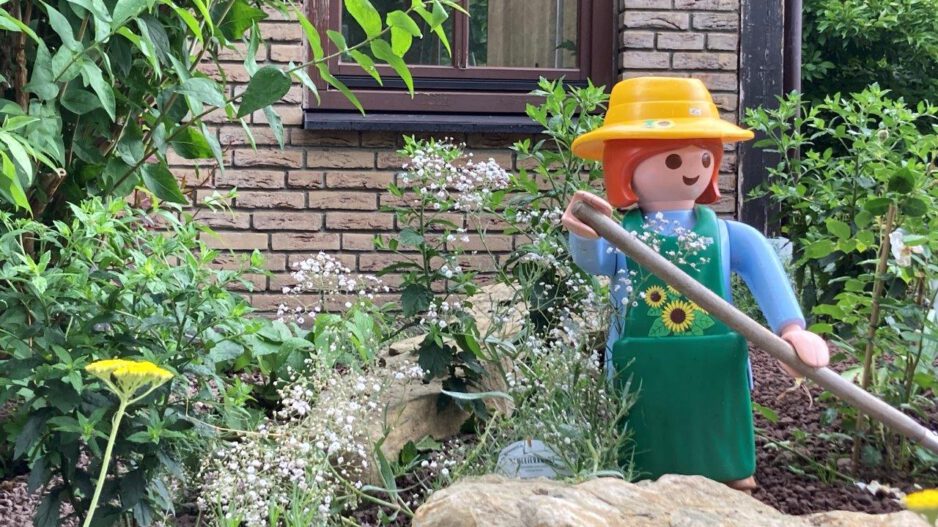 Eine große Spielfigur im Stil einer Gärtnerin mit gelbem Hut, grüner Schürze und langem Gartenwerkzeug steht zwischen Blumen, Sträuchern und Natursteinen in einem liebevoll angelegten Vorgarten. Im Hintergrund sind die Klinkerfassade und ein Fenster eines Wohnhauses zu sehen. Die Szene vermittelt den Eindruck eines gepflegten, naturnah gestalteten Gartens mit dekorativer Figur.