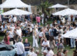 Zahlreiche Besucherinnen und Besucher genießen ein Sommerfest im Freien auf einer Wiese mit Bierbänken, Stehtischen und großen Sonnenschirmen. Die Menschen sitzen, stehen und unterhalten sich in entspannter Atmosphäre, während im Hintergrund ein historisches Gebäude und ein Festzelt zu sehen sind. Eine Palme in der Mitte des Geländes verleiht der Veranstaltung ein sommerliches Ambiente.