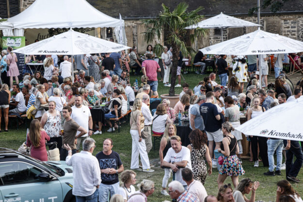 Foto: Dennis Echtermann Zahlreiche Besucherinnen und Besucher genießen ein Sommerfest im Freien auf einer Wiese mit Bierbänken, Stehtischen und großen Sonnenschirmen. Die Menschen sitzen, stehen und unterhalten sich in entspannter Atmosphäre, während im Hintergrund ein historisches Gebäude und ein Festzelt zu sehen sind. Eine Palme in der Mitte des Geländes verleiht der Veranstaltung ein sommerliches Ambiente.