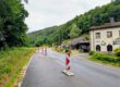 Mehrere rot-weiße Leitbaken verengen die Fahrbahn einer Landstraße und weisen auf eine Baustelle hin. Die Straße führt durch ein bewaldetes Tal und verläuft an einem Wohnhaus sowie weiteren Gebäuden vorbei. Der nasse Asphalt und der bedeckte Himmel lassen auf regnerisches Wetter schließen, während die provisorische Verkehrsführung den Verkehr sicher an der Baustelle vorbeileitet.
