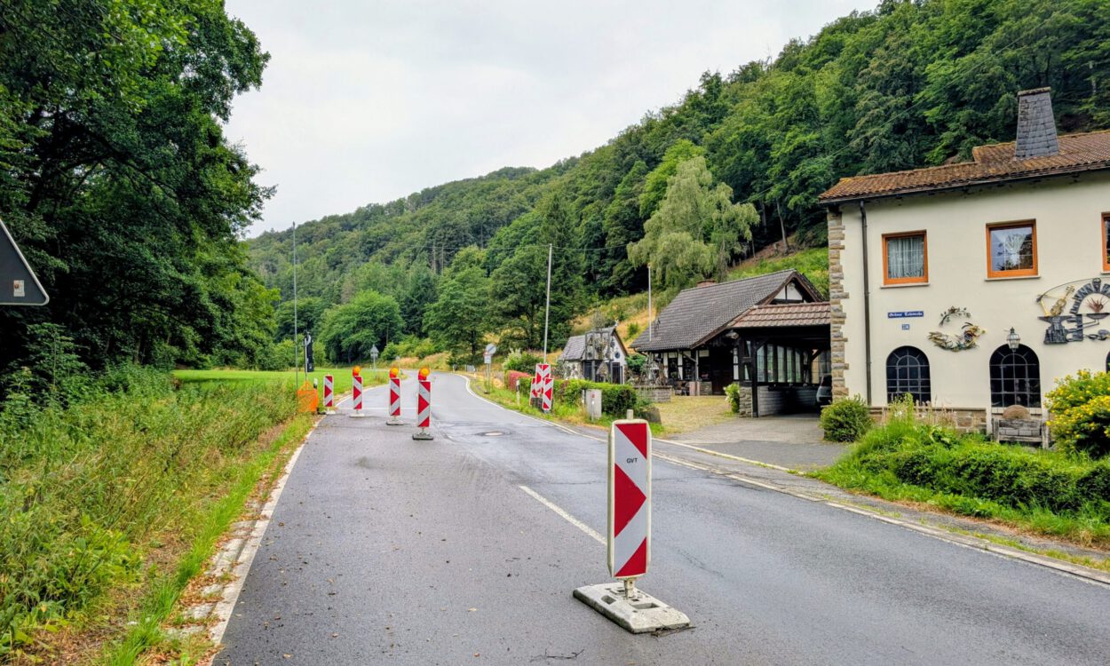 Mehrere rot-weiße Leitbaken verengen die Fahrbahn einer Landstraße und weisen auf eine Baustelle hin. Die Straße führt durch ein bewaldetes Tal und verläuft an einem Wohnhaus sowie weiteren Gebäuden vorbei. Der nasse Asphalt und der bedeckte Himmel lassen auf regnerisches Wetter schließen, während die provisorische Verkehrsführung den Verkehr sicher an der Baustelle vorbeileitet.