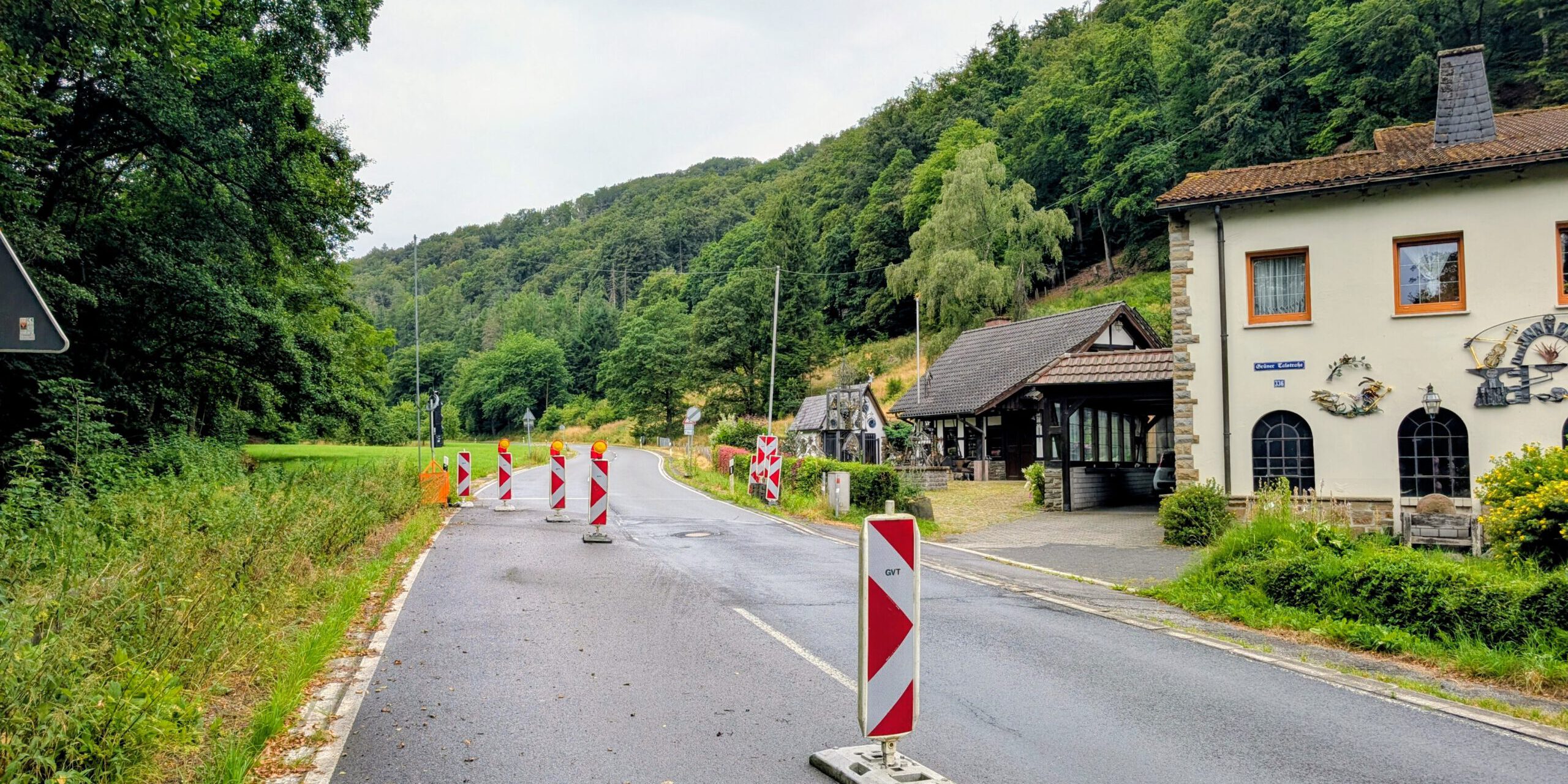Mehrere rot-weiße Leitbaken verengen die Fahrbahn einer Landstraße und weisen auf eine Baustelle hin. Die Straße führt durch ein bewaldetes Tal und verläuft an einem Wohnhaus sowie weiteren Gebäuden vorbei. Der nasse Asphalt und der bedeckte Himmel lassen auf regnerisches Wetter schließen, während die provisorische Verkehrsführung den Verkehr sicher an der Baustelle vorbeileitet.
