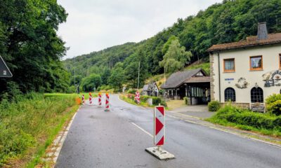 Mehrere rot-weiße Leitbaken verengen die Fahrbahn einer Landstraße und weisen auf eine Baustelle hin. Die Straße führt durch ein bewaldetes Tal und verläuft an einem Wohnhaus sowie weiteren Gebäuden vorbei. Der nasse Asphalt und der bedeckte Himmel lassen auf regnerisches Wetter schließen, während die provisorische Verkehrsführung den Verkehr sicher an der Baustelle vorbeileitet.