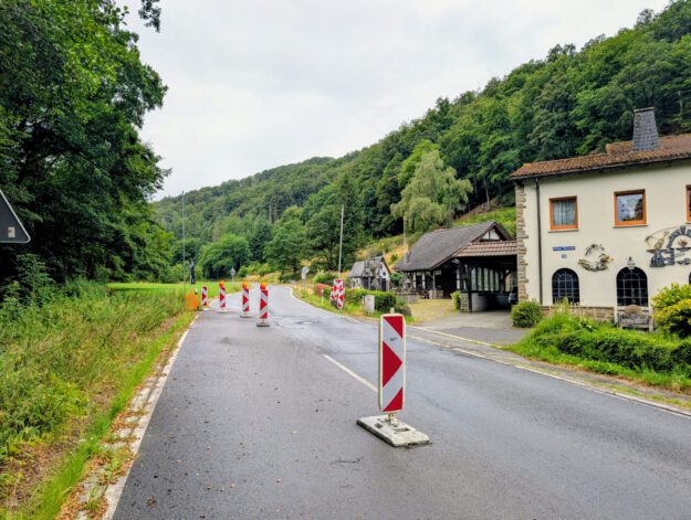 Mehrere rot-weiße Leitbaken verengen die Fahrbahn einer Landstraße und weisen auf eine Baustelle hin. Die Straße führt durch ein bewaldetes Tal und verläuft an einem Wohnhaus sowie weiteren Gebäuden vorbei. Der nasse Asphalt und der bedeckte Himmel lassen auf regnerisches Wetter schließen, während die provisorische Verkehrsführung den Verkehr sicher an der Baustelle vorbeileitet.