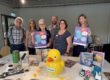 Sechs Personen stehen hinter einem Tisch in einer Bibliothek und präsentieren Plakate für den „SLC SommerLeseClub“. Auf dem Tisch liegen Kinder- und Jugendbücher, eine große gelbe Ente mit der Aufschrift „Mama Berta“, mehrere kleine Badeenten sowie Bastelarbeiten und Werbematerialien. Die Aufnahme vermittelt den Eindruck einer gemeinsamen Auftaktveranstaltung zur Leseförderung und lädt zur Teilnahme am Sommerleseclub ein.