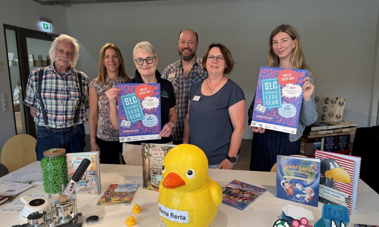 Sechs Personen stehen hinter einem Tisch in einer Bibliothek und präsentieren Plakate für den „SLC SommerLeseClub“. Auf dem Tisch liegen Kinder- und Jugendbücher, eine große gelbe Ente mit der Aufschrift „Mama Berta“, mehrere kleine Badeenten sowie Bastelarbeiten und Werbematerialien. Die Aufnahme vermittelt den Eindruck einer gemeinsamen Auftaktveranstaltung zur Leseförderung und lädt zur Teilnahme am Sommerleseclub ein.