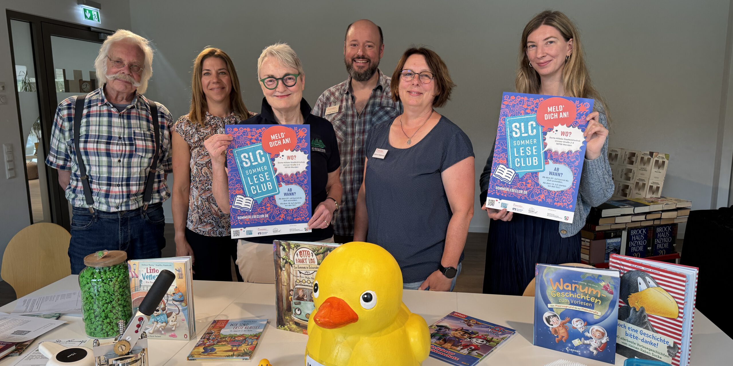 Sechs Personen stehen hinter einem Tisch in einer Bibliothek und präsentieren Plakate für den „SLC SommerLeseClub“. Auf dem Tisch liegen Kinder- und Jugendbücher, eine große gelbe Ente mit der Aufschrift „Mama Berta“, mehrere kleine Badeenten sowie Bastelarbeiten und Werbematerialien. Die Aufnahme vermittelt den Eindruck einer gemeinsamen Auftaktveranstaltung zur Leseförderung und lädt zur Teilnahme am Sommerleseclub ein.