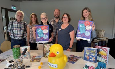 Sechs Personen stehen hinter einem Tisch in einer Bibliothek und präsentieren Plakate für den „SLC SommerLeseClub“. Auf dem Tisch liegen Kinder- und Jugendbücher, eine große gelbe Ente mit der Aufschrift „Mama Berta“, mehrere kleine Badeenten sowie Bastelarbeiten und Werbematerialien. Die Aufnahme vermittelt den Eindruck einer gemeinsamen Auftaktveranstaltung zur Leseförderung und lädt zur Teilnahme am Sommerleseclub ein.